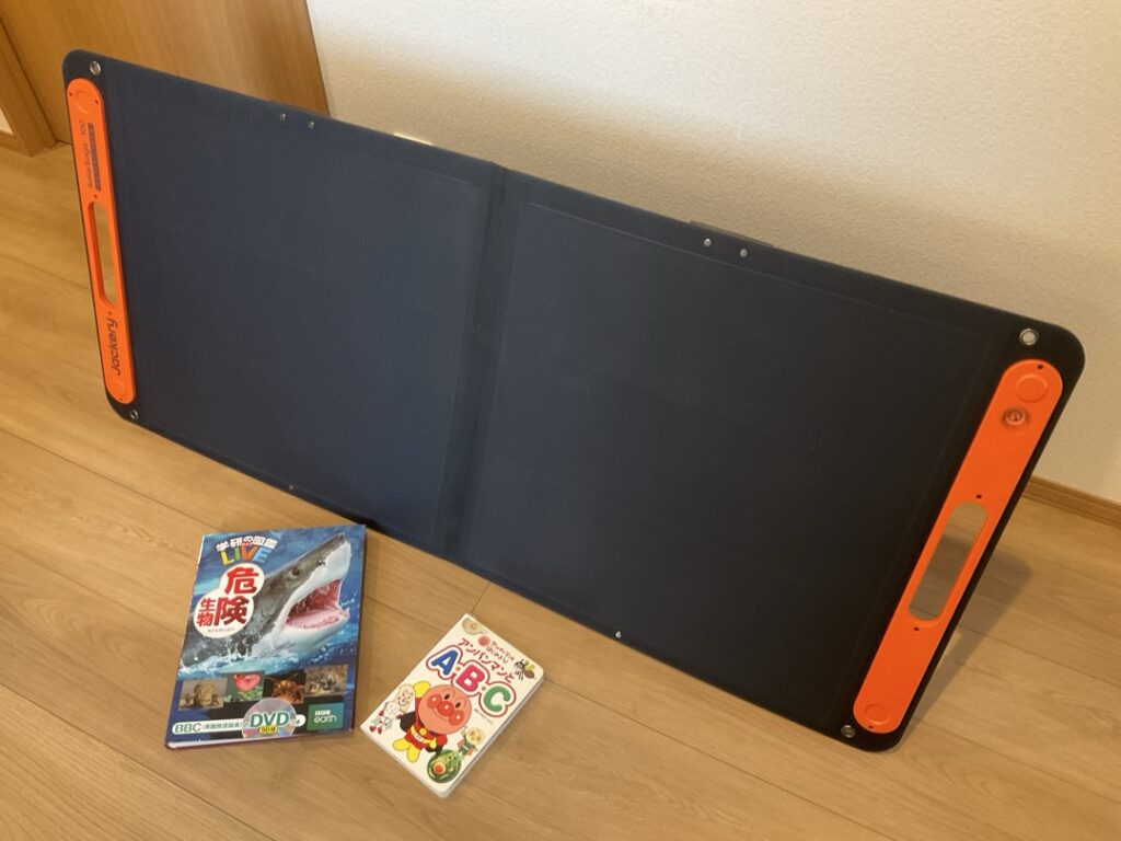 Jackery SolarSaga100と子どもの図鑑を並べて大きさを比較した写真。家庭で使う物と並べることで、ソーラーパネルの実際のサイズ感が分かりやすい構図。
